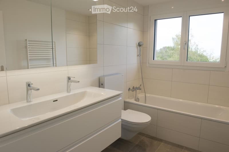 Modern bathroom with white tiles, a large sink with dual faucets, a bathtub, a toilet, and a large mirror above the sink. There is a radiator and a window letting in natural light.
