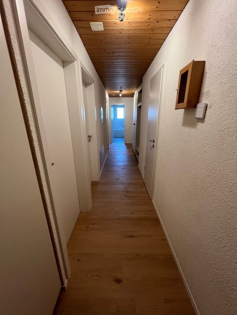 A long, narrow hallway with wooden flooring, white walls, and several closed doors on both sides. The ceiling features wooden paneling and recessed lighting.
