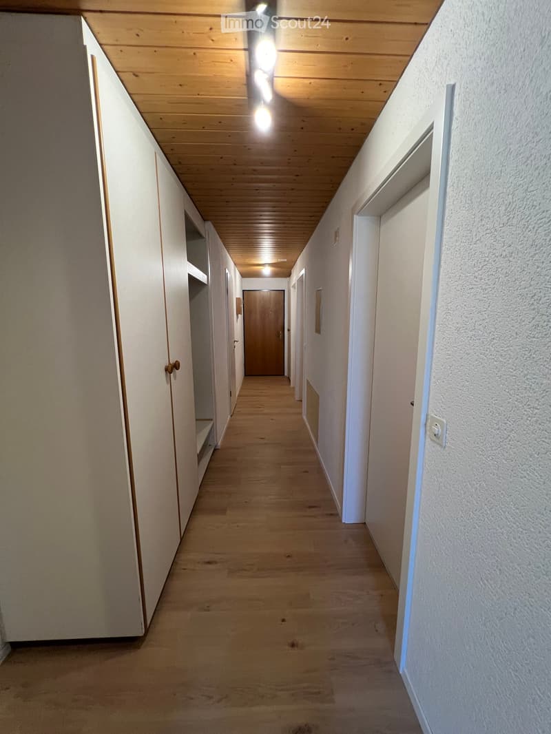 A long, narrow hallway with wooden flooring and white walls, featuring several doors and built-in shelving on one side.