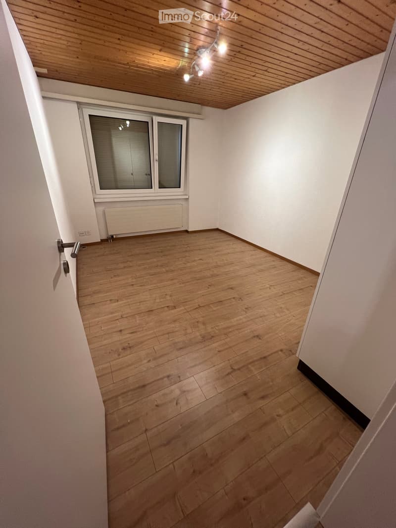 Empty room with wooden flooring, white walls, and a window with blinds. The ceiling has wooden paneling and a light fixture.