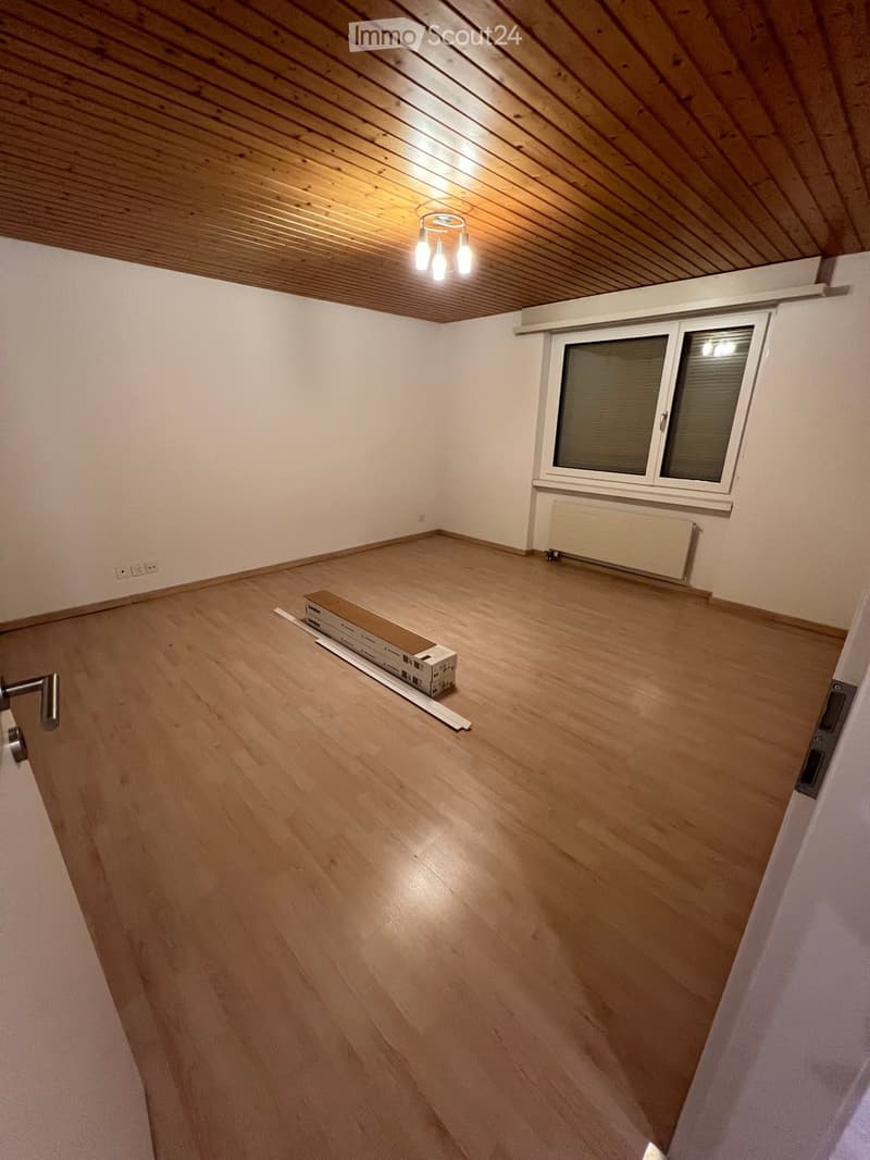 Empty room with wooden flooring, white walls, and a slanted wooden ceiling. A single window with blinds is visible, along with a radiator below it. A small cardboard box and a white plank are placed on the floor.