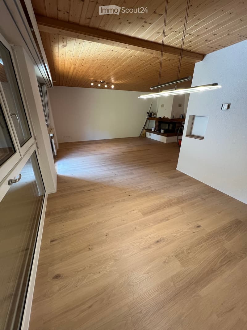 Empty room with wooden flooring, white walls, and a wooden ceiling. Features include a large window with blinds, a hanging light fixture, and a small built-in shelf with a few items.