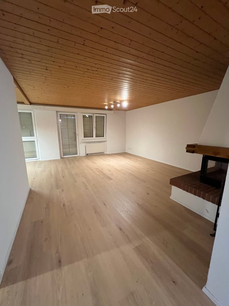 Empty room with wooden flooring, white walls, and a slanted wooden ceiling. Features include a window with blinds, a radiator, and a small wooden platform with a brick base.