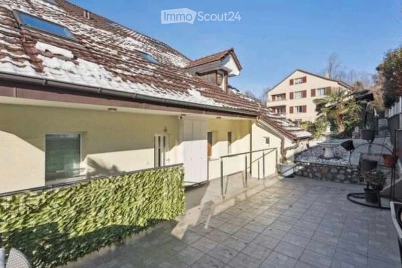 Courtyard area with tiled flooring, low green hedge along a railing, small fountain, and potted plants. Adjacent building has a white door and window, with snow on the roof of the main house.