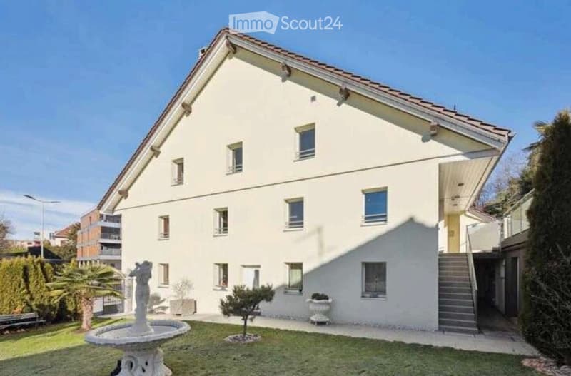 A modern two-story house with white exterior walls, multiple windows, and a sloped roof. The house features a small garden area with a fountain, shrubs, and a staircase leading to the entrance.