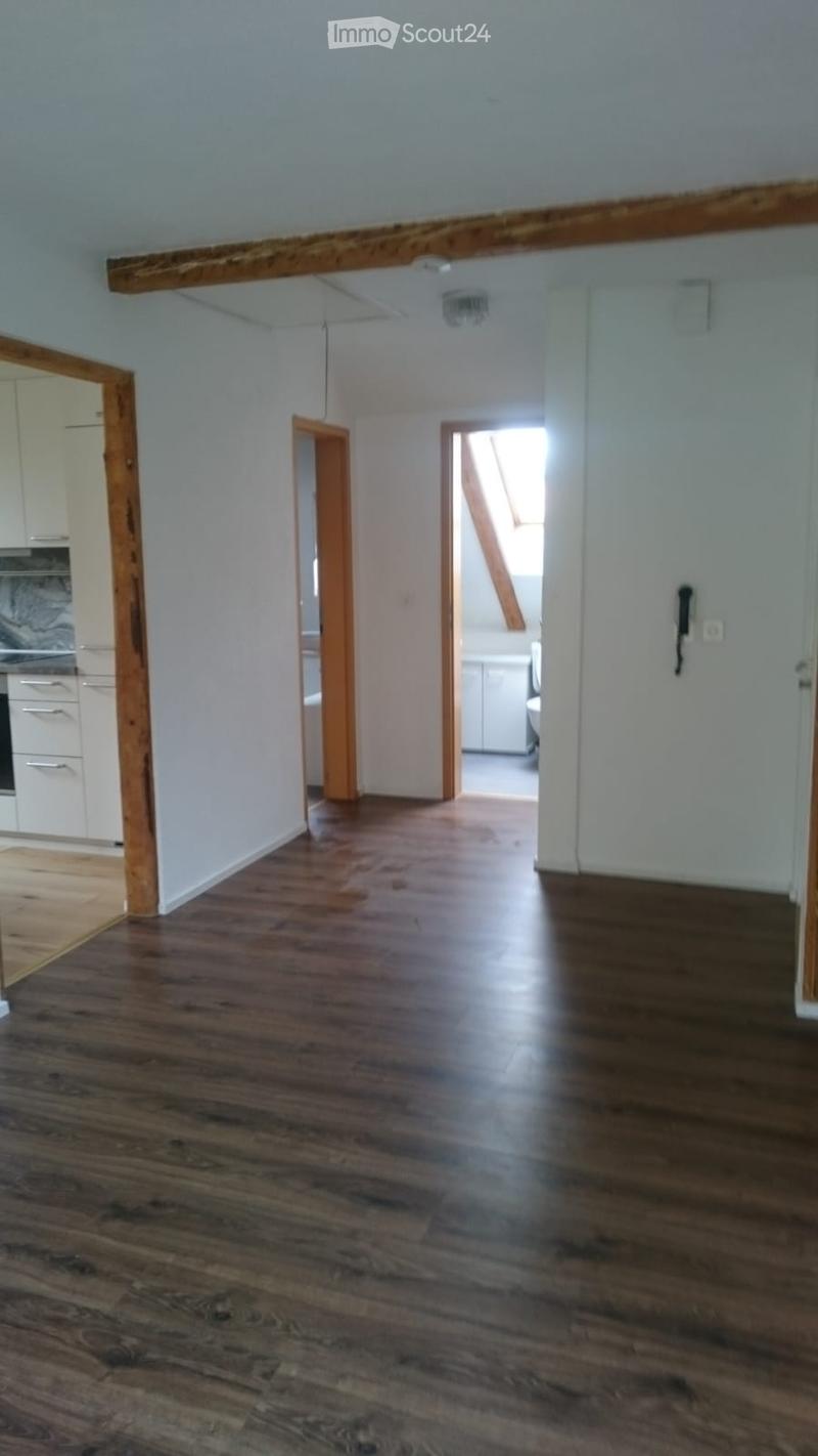 Empty hallway with wooden flooring, white walls, and exposed wooden beams on the ceiling. The hallway leads to other rooms, including a kitchen and a staircase area.