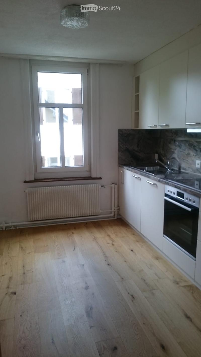 Empty kitchen with light wooden flooring, white cabinets, and a window letting in natural light. No appliances or furniture are present.