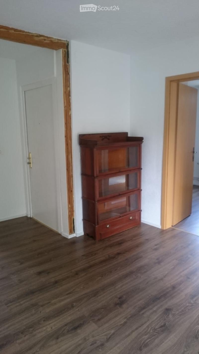 A small, unfurnished room with wooden flooring and white walls. A wooden cabinet with glass doors and drawers is positioned against one wall. Two doors are visible, one on each side of the room.