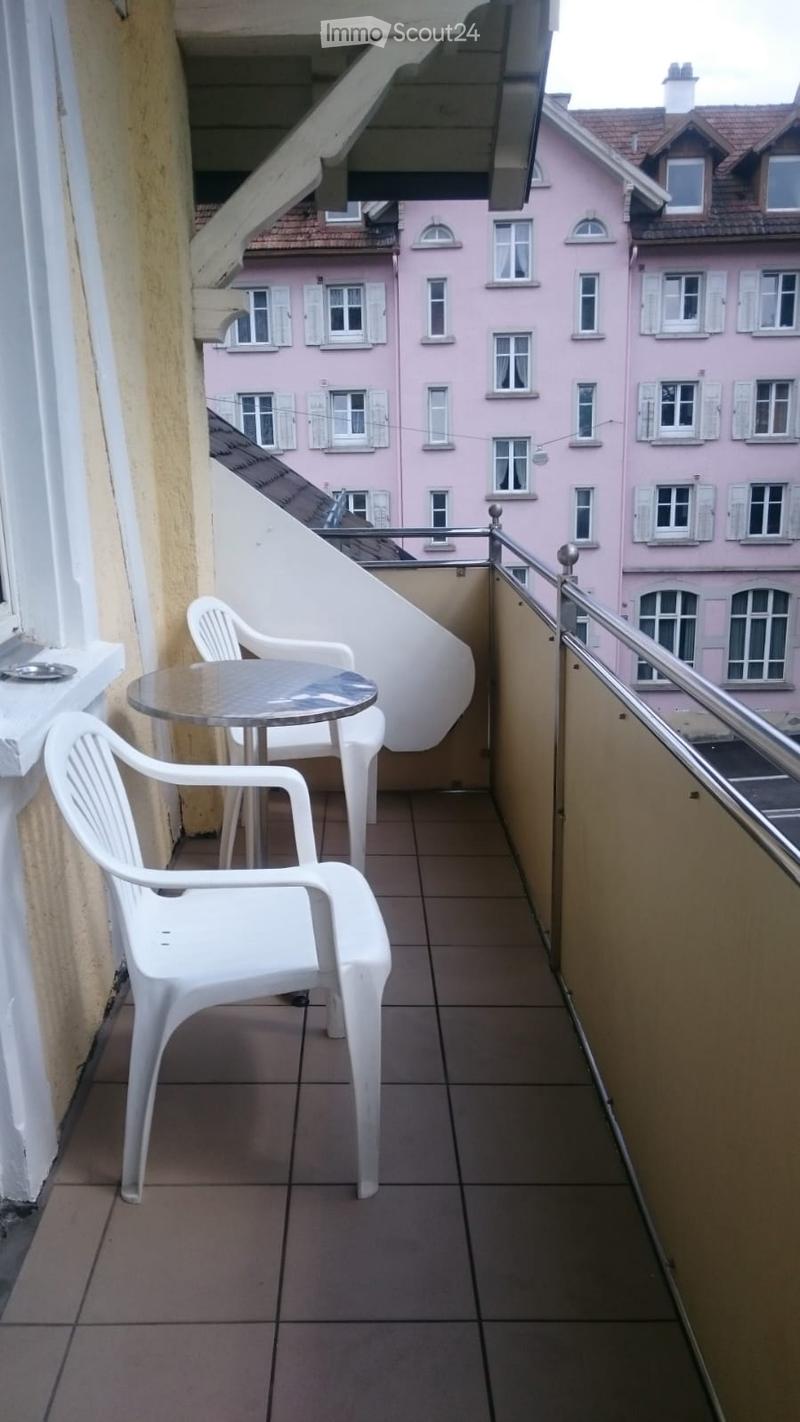 Balcony with tiled floor, white plastic chairs, and a small round table, overlooking a pink residential building with multiple windows.