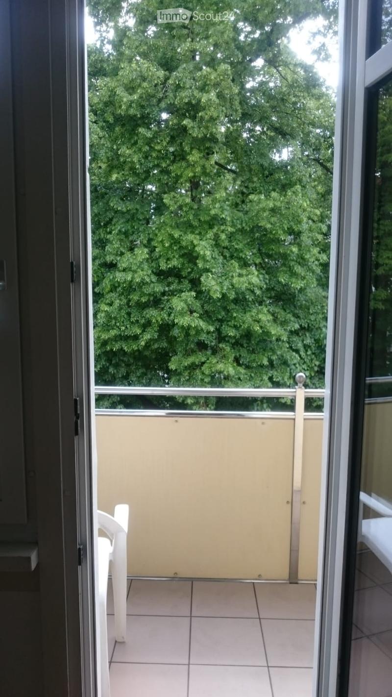 Balcony with tiled floor, white plastic chair, glass railing, and view of green trees.