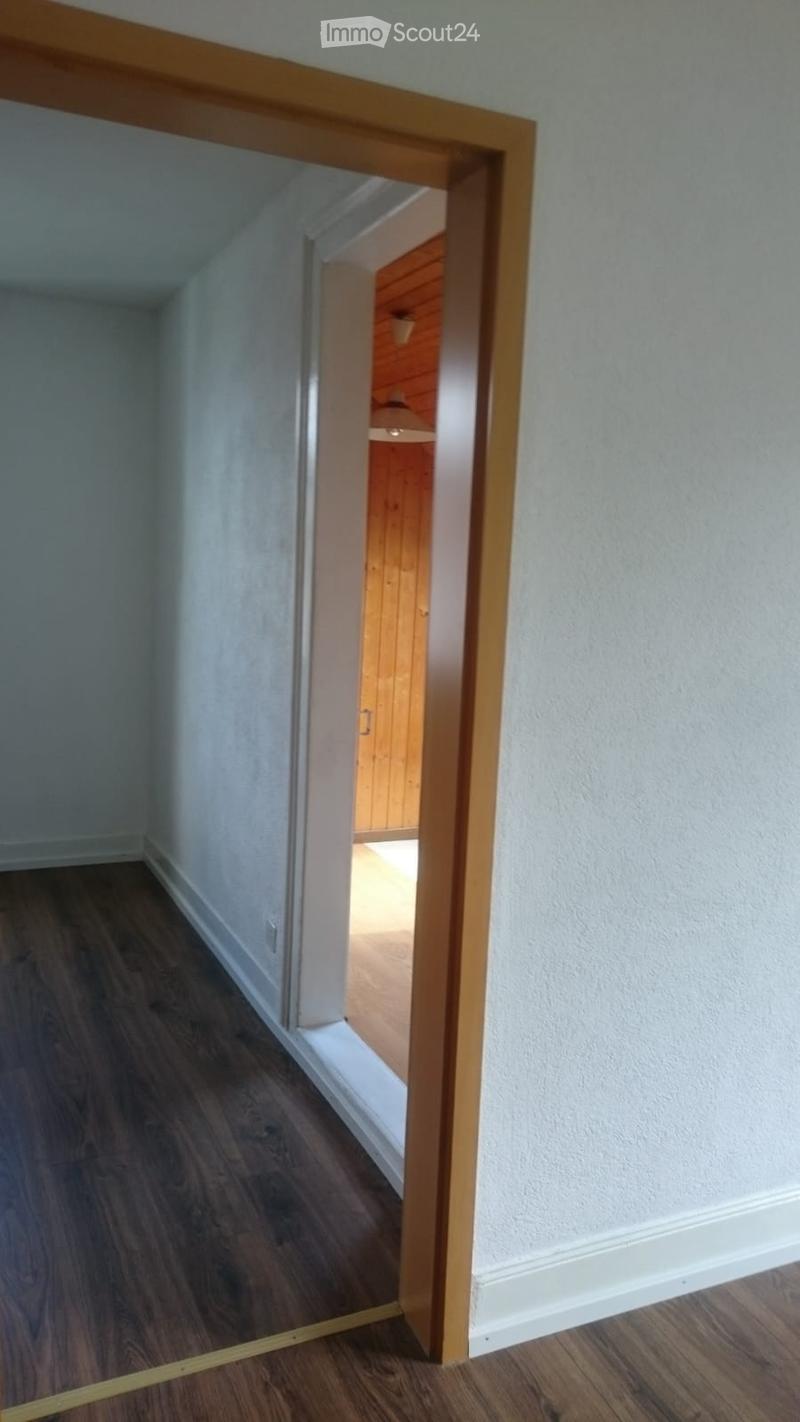 Empty hallway with light-colored walls, wooden flooring, and an open doorway leading to another room with wooden paneling.