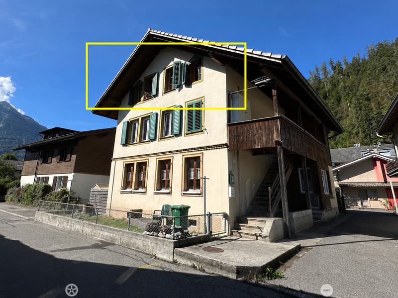 Two-story house with beige walls, green shutters, and a wooden balcony. The house is situated in a scenic area with mountains in the background.