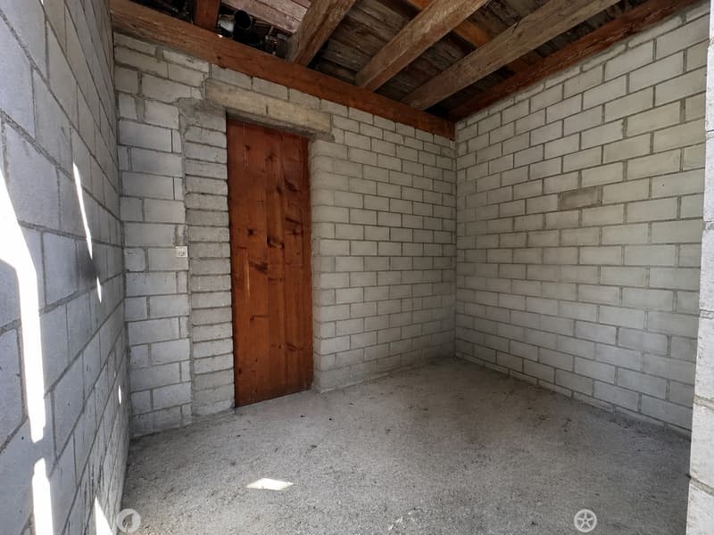 Empty room with exposed brick walls, wooden door, and wooden ceiling beams. Concrete floor, no furniture or equipment.