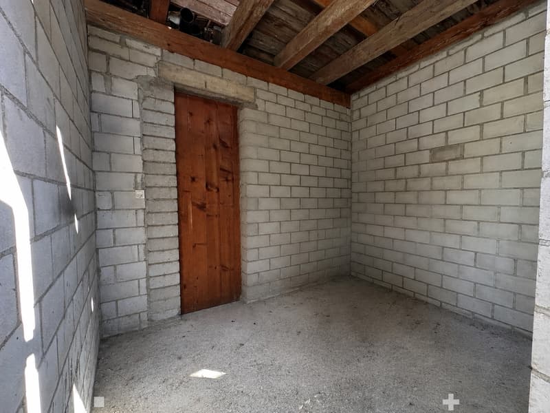 Empty room with exposed brick walls, wooden door, and wooden ceiling beams. Concrete floor, no furniture or equipment.
