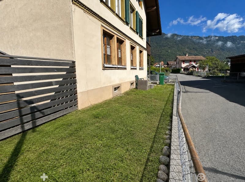 A small, well-maintained garden area with a wooden fence, green lawn, and a view of a residential street and distant mountains.