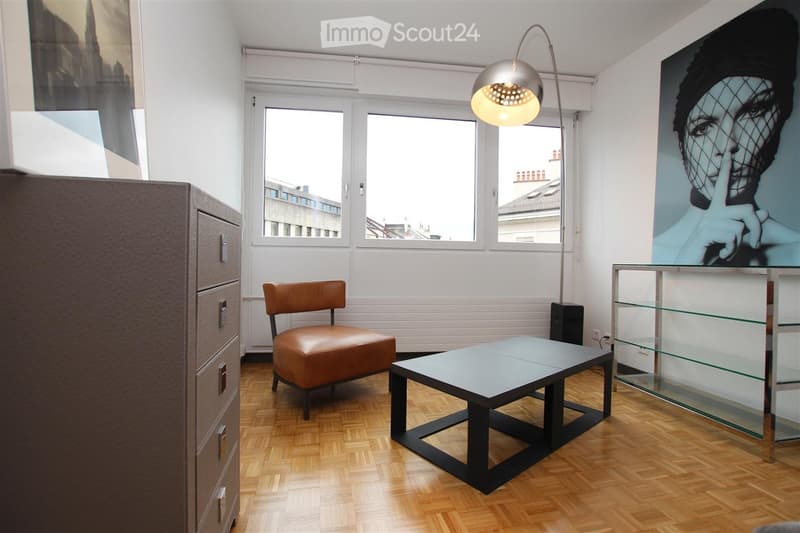 Modern living room with wooden flooring, large windows, a brown leather chair, a square glass-top table, a tall floor lamp, and a glass display cabinet.