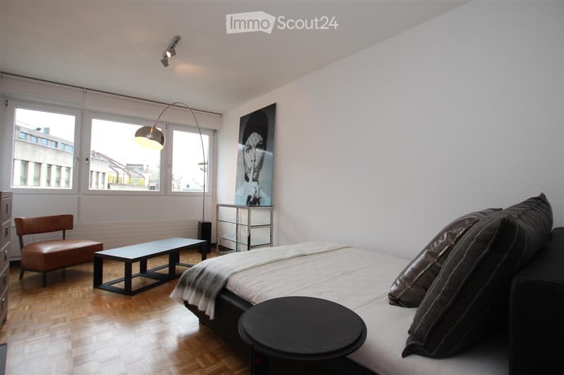 Modern, well-lit room with a sofa, a small table, a chair, a floor lamp, and a picture frame on the wall. The room features large windows and a wooden floor.