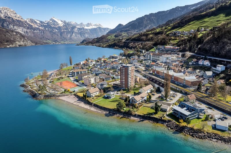 Aerial view of a town nestled by a lake with surrounding mountains, featuring residential buildings, a beach, and a tennis court.