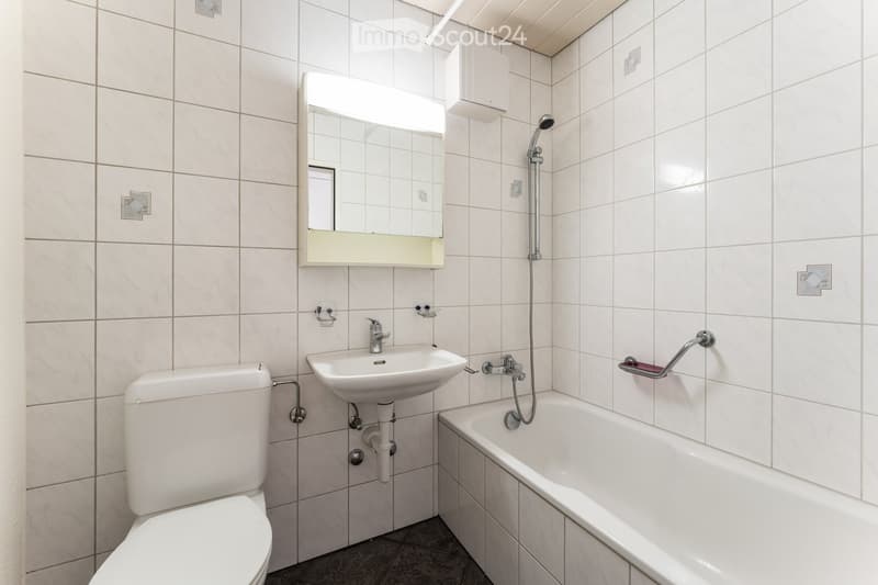 Modern bathroom with white tiled walls and floor, featuring a toilet, sink, bathtub, and mirror with built-in lighting.