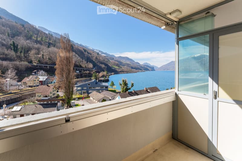 Balcony with expansive view of a lake and surrounding mountains, featuring modern glass doors and a clean, minimalist design.