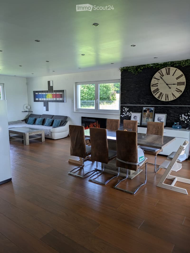 Modern living and dining area with wooden flooring, a white sectional sofa, a glass dining table with six chairs, a fireplace with a large clock above it, and several picture frames on the wall.