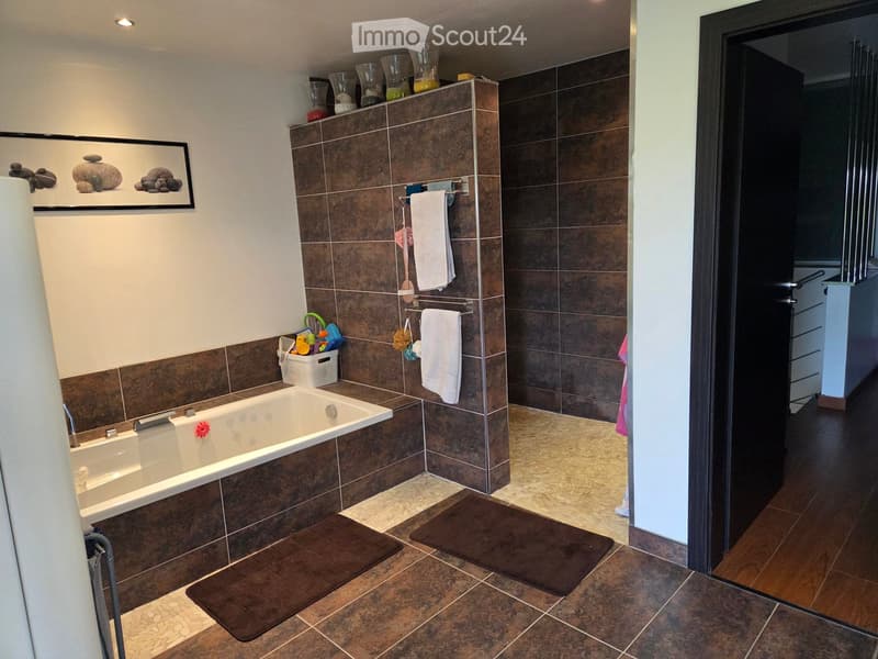 Bathroom with a bathtub, towel rack, towels, bathroom mats, and a framed picture on the wall. The room features brown tiles and a white countertop.