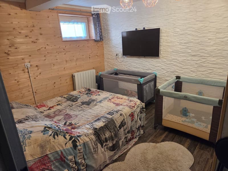 Cozy bedroom with wooden walls, a large bed with a colorful blanket, two baby cribs, a round rug, a radiator, and a mounted TV.