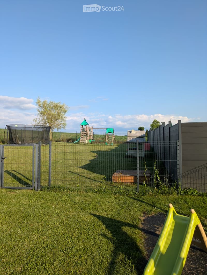 A fenced backyard with a grassy lawn, a small play structure, a trampoline, and a slide visible in the foreground.