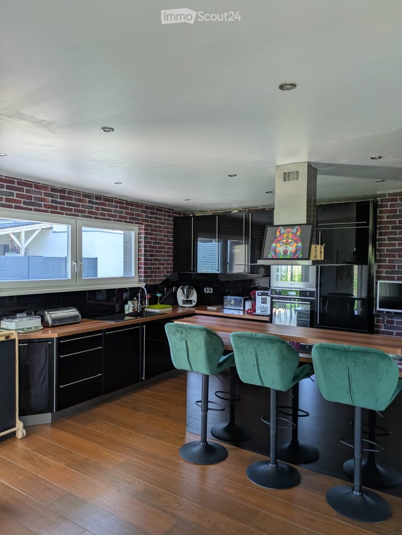 Modern kitchen with wooden countertops, black cabinets, and three green stools at the island. Features a sink, faucet, oven, refrigerator, microwave, coffee maker, and a bread maker on the counter.