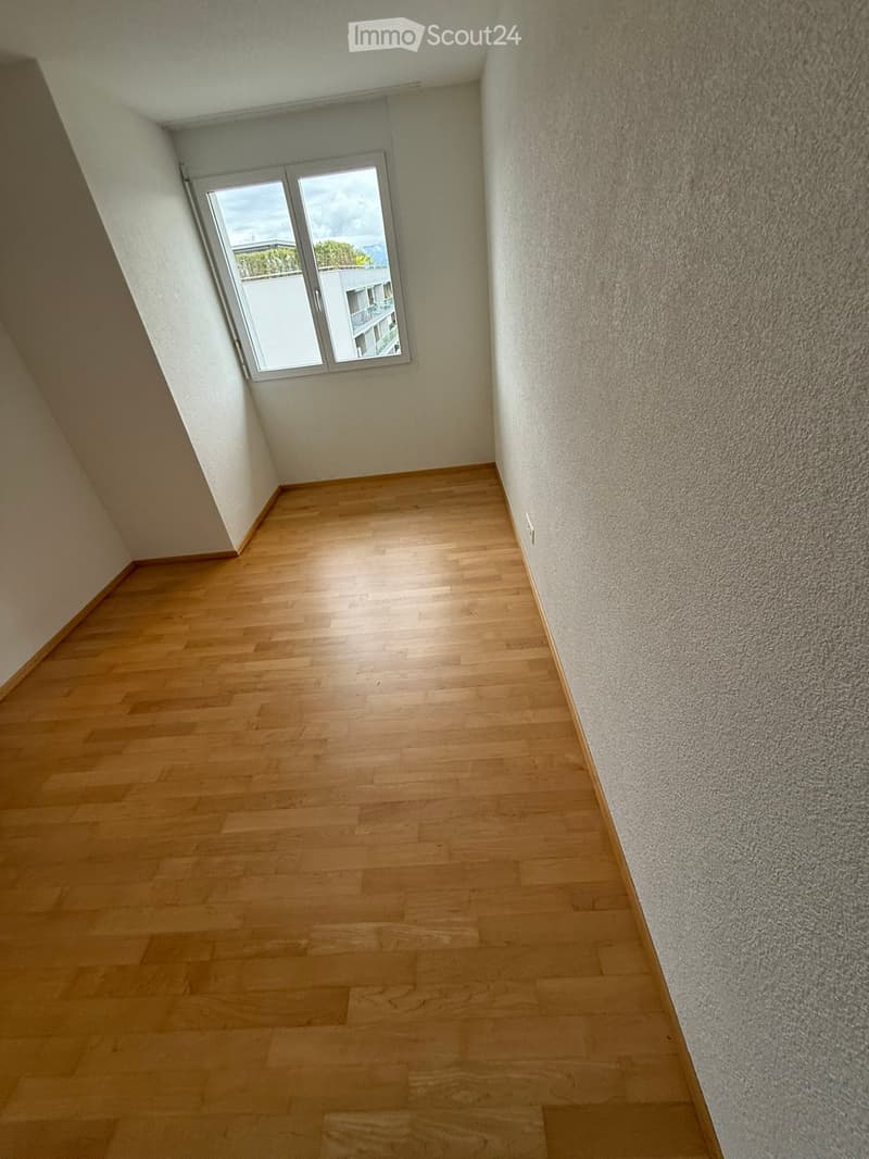 Empty hallway with wooden flooring and a window at the end, showing a view of a building and greenery outside.