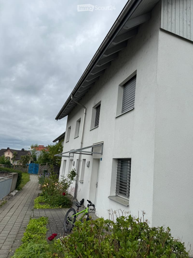 Two-story white house with multiple windows, a covered entrance, and a small garden area with plants and a bicycle.