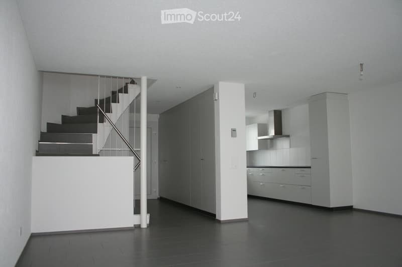 Empty room with white walls, gray floor, white kitchen cabinets, white ceiling, metal handrail, and a staircase.