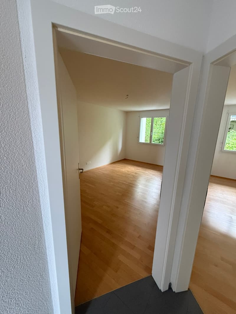Empty room with wooden flooring, white walls, and large windows letting in natural light. The room is unfurnished and appears to be part of a larger residential space.