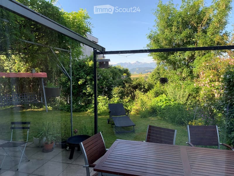 Garden with a wooden table and chairs, sun lounger, swing, fire pit, and potted plant, surrounded by lush greenery and a clear view of mountains.