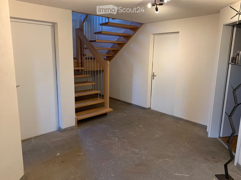 Empty hallway with a wooden staircase leading upwards, white walls, and a concrete floor.