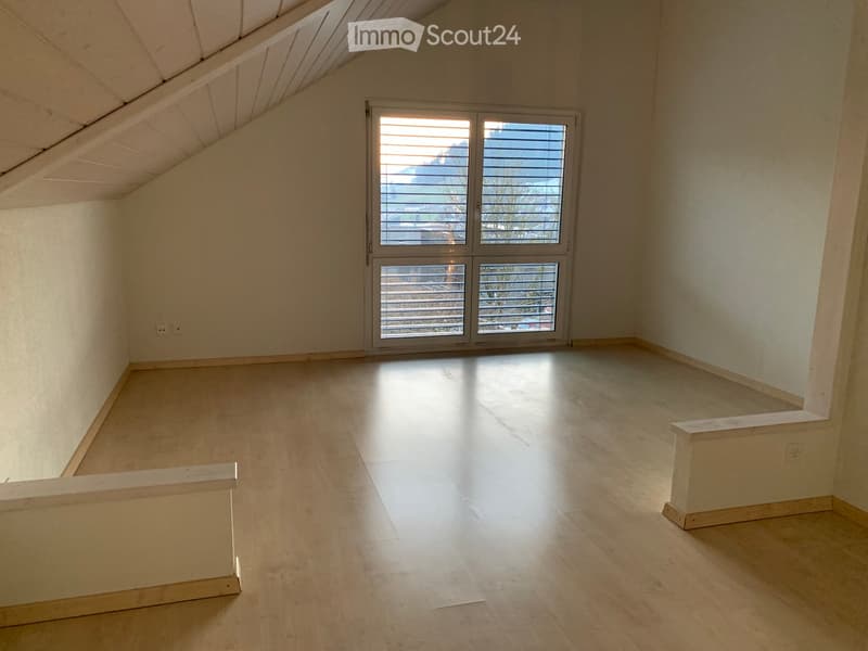 Empty attic room with wooden flooring, white walls, and a large window with blinds offering a view of the outdoors.
