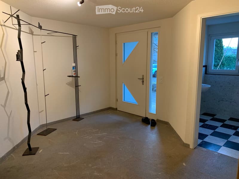 Empty hallway with tiled floor, white walls, a door with glass panels, and a small shelf with a bottle on it. A pair of shoes is placed near the door.