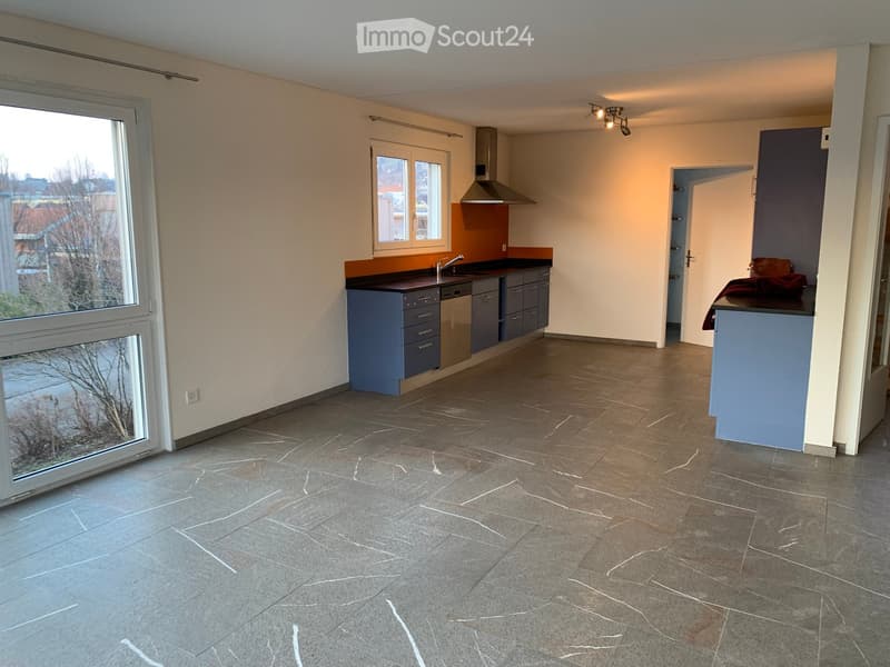 Open-plan kitchen and living area with grey tiled flooring, blue cabinets, stainless steel sink, dishwasher, and a bar area with a stool.