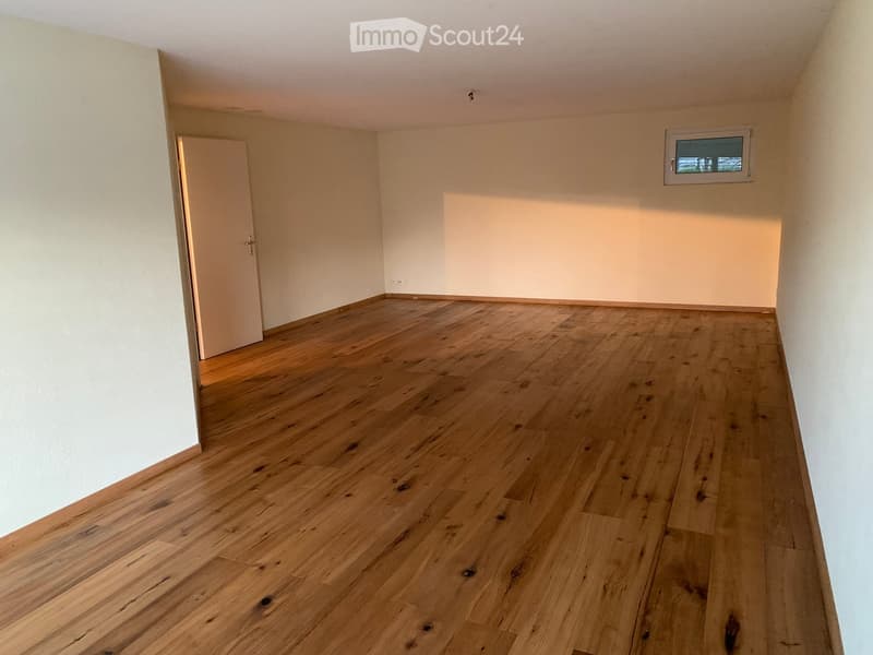 Empty room with light wooden flooring and white walls, featuring a small window and a door leading to another space.