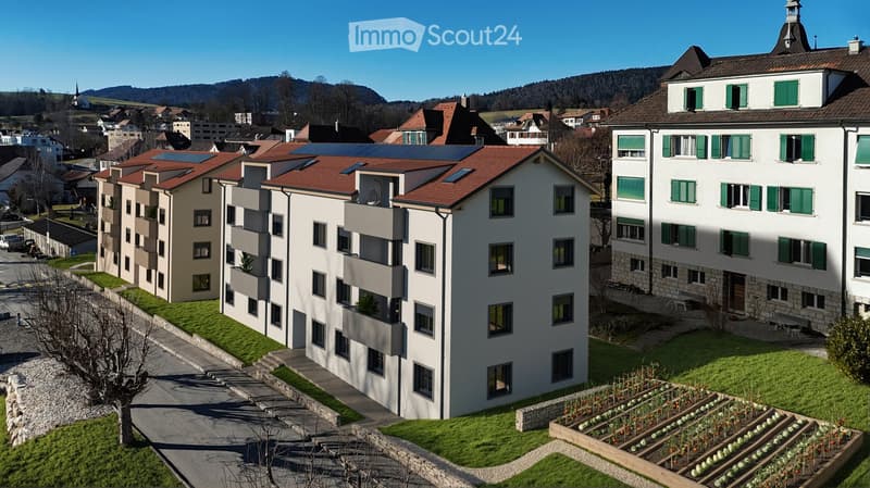 Modern multi-story residential building with solar panels on the roof, surrounded by a landscaped garden area with raised beds and pathways.