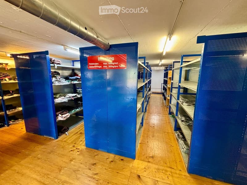 A long, narrow hallway with wooden flooring and blue shelving units on both sides, containing folded clothes and other stored items. The hallway is well-lit with fluorescent lighting and has a sign indicating weight limits for storage.