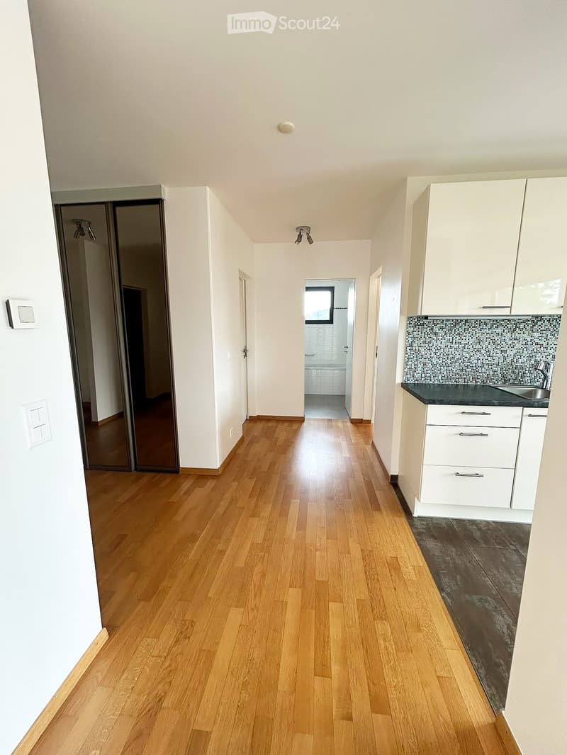 Empty hallway with wooden flooring, white walls, and a small kitchen area visible on the right side.