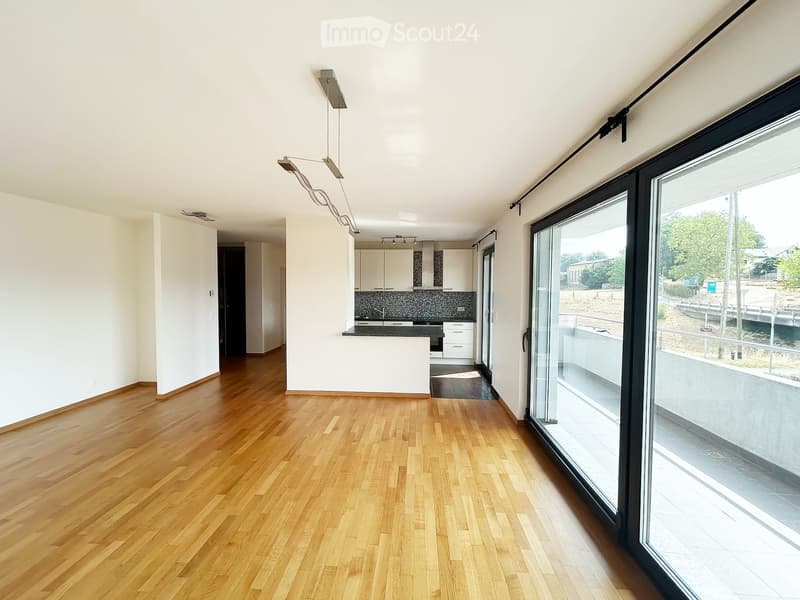 Bright, unfurnished living space with wooden flooring, large glass sliding doors leading to a balcony, and a modern kitchen area visible in the background.