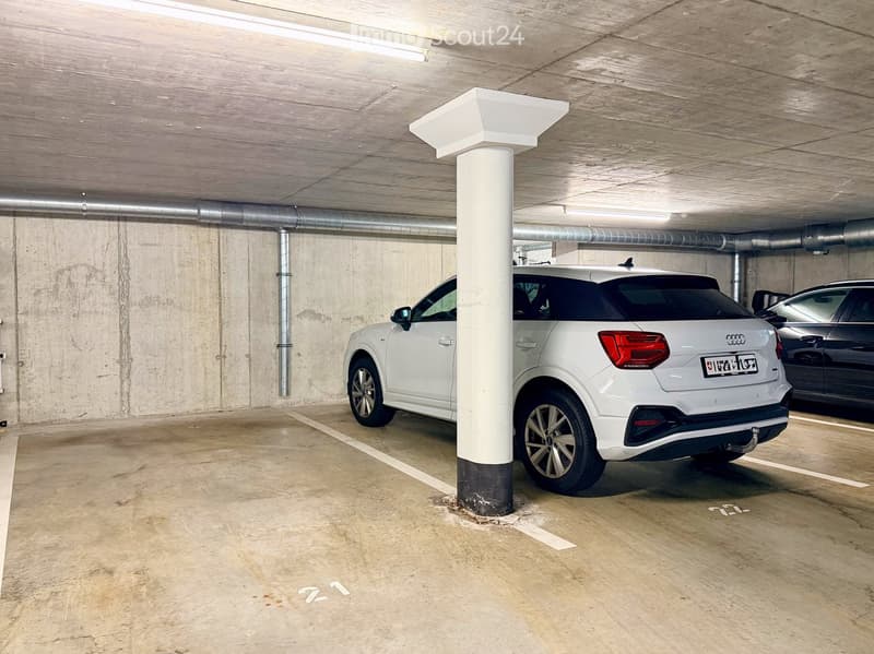 Underground parking area with concrete floors and walls, featuring a white Audi SUV and a dark-colored car parked in designated spots.