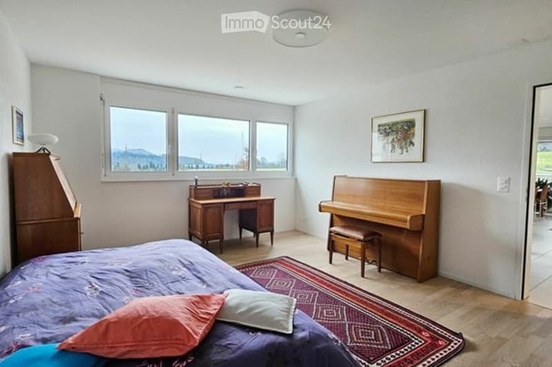Bright bedroom with large windows offering a scenic view, featuring a bed with colorful pillows, a wooden desk, a piano, and a wardrobe.