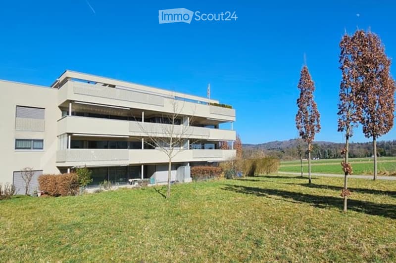 Modern multi-story apartment building with balconies, surrounded by a grassy lawn and trees under a clear blue sky.