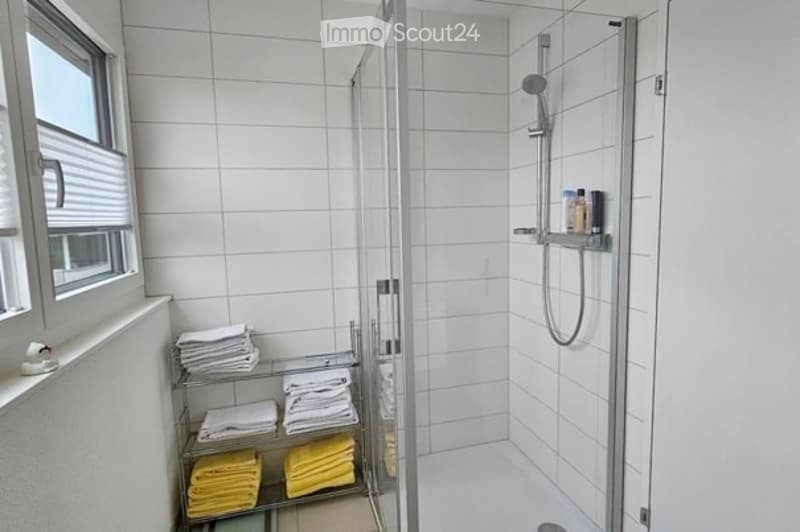 Modern bathroom with white tiled walls and floor, a glass-enclosed shower stall, and a towel rack holding neatly folded towels.