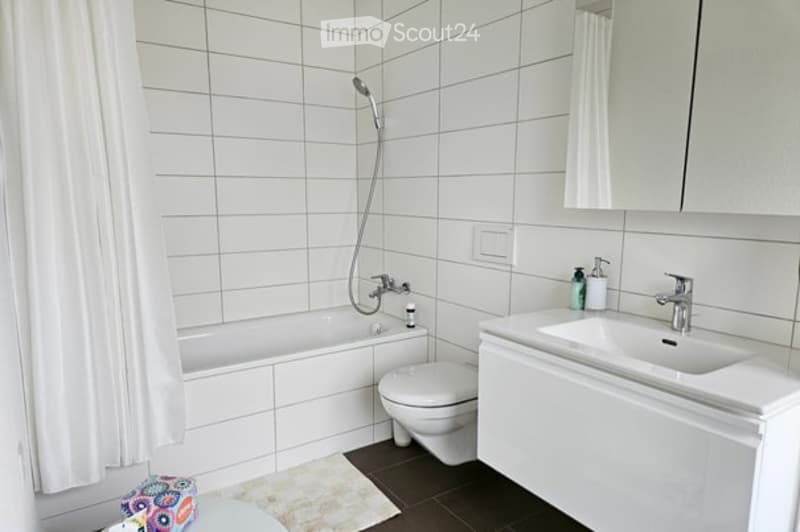 Modern bathroom with white tiles, a bathtub, toilet, sink, mirror, and a shower hose attached to the wall.