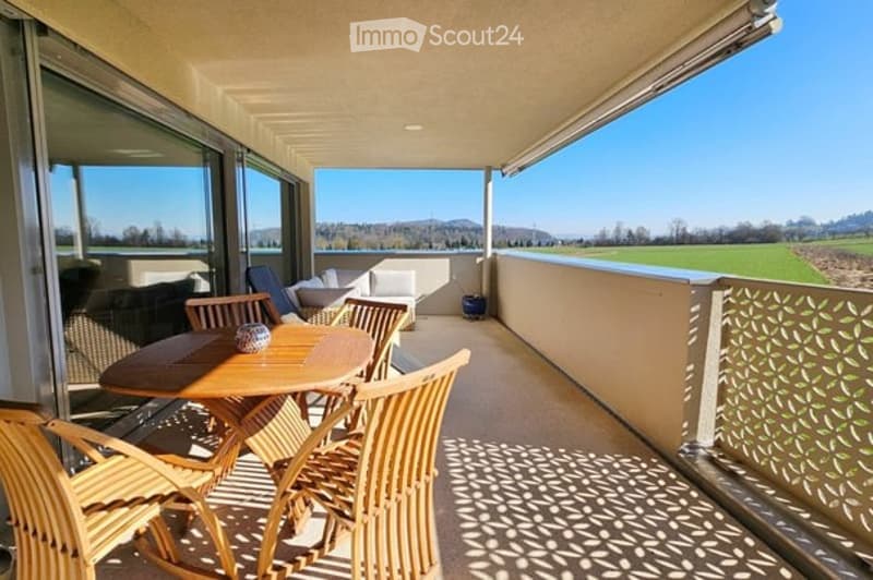 Spacious balcony with wooden dining table and chairs, white sofa, and scenic countryside view.