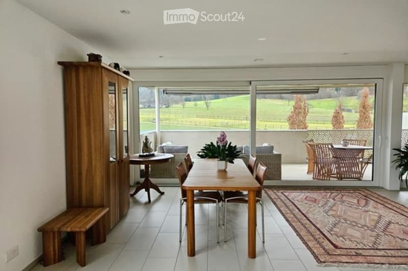 Bright dining area with large windows overlooking a green field, featuring a wooden dining table, chairs, a bench, side table, vase with flowers, and a patterned rug.